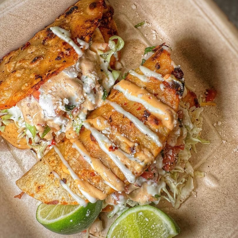 Two Acapela Modern Mex tacos with white and orange sauce, lettuce, and lime wedges.