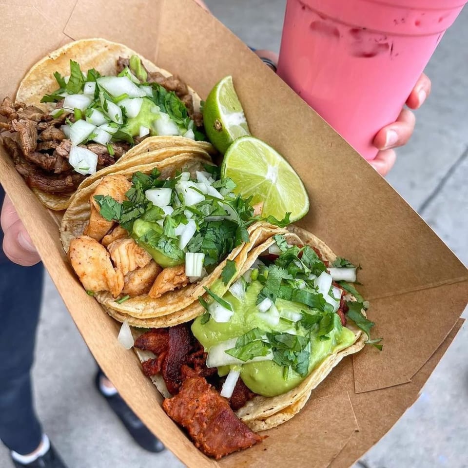 Three Acapela Modern Mex tacos with various fillings, cilantro, onions, and lime wedges in a tray.